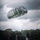 A military paratrooper is captured at the moment of landing in an open green field, suspended just above the ground beneath a large round military-style parachute in dark olive/gray coloring. The scene takes place against a dramatic overcast sky with dense tree lines in the background. The image appears to document a military airborne training or operational jump exercise.
