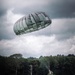 A military paratrooper is captured at the moment of landing in an open green field, suspended just above the ground beneath a large round military-style parachute in dark olive/gray coloring. The scene takes place against a dramatic overcast sky with dense tree lines in the background. The image appears to document a military airborne training or operational jump exercise.