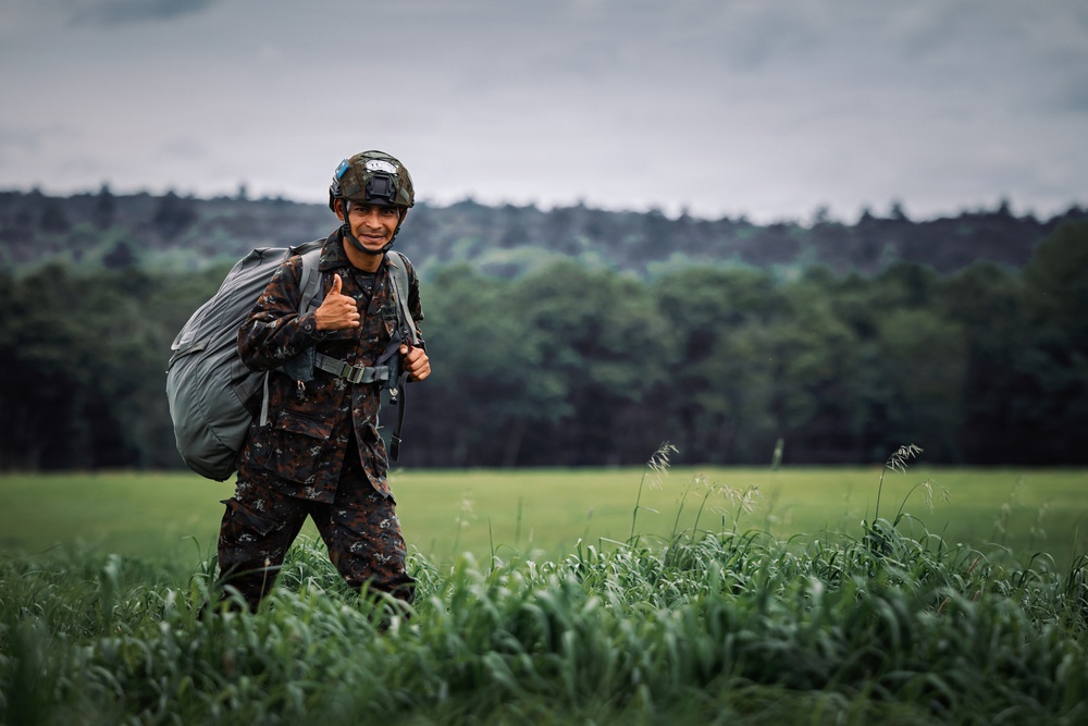A smiling soldier in camouflage uniform and tactical helmet gives a thumbs-up gesture while standing in a green field. He is carrying what appears to be a packed parachute or large equipment bag on his back. The background features a treeline and overcast sky, suggesting an outdoor military exercise or post-jump landing zone.