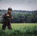 A smiling soldier in camouflage uniform and tactical helmet gives a thumbs-up gesture while standing in a green field. He is carrying what appears to be a packed parachute or large equipment bag on his back. The background features a treeline and overcast sky, suggesting an outdoor military exercise or post-jump landing zone.