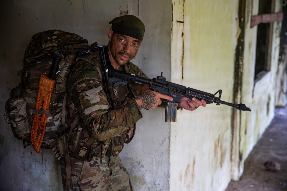 A male individual in full camouflage military-style gear is positioned against a weathered interior wall, holding a rifle in a tactical stance. He is wearing a dark green beret, carrying a large tactical backpack, and has visible tattoos on his forearms. The setting appears to be a dilapidated or abandoned building with peeling paint and aged walls.
