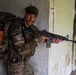 A male individual in full camouflage military-style gear is positioned against a weathered interior wall, holding a rifle in a tactical stance. He is wearing a dark green beret, carrying a large tactical backpack, and has visible tattoos on his forearms. The setting appears to be a dilapidated or abandoned building with peeling paint and aged walls.