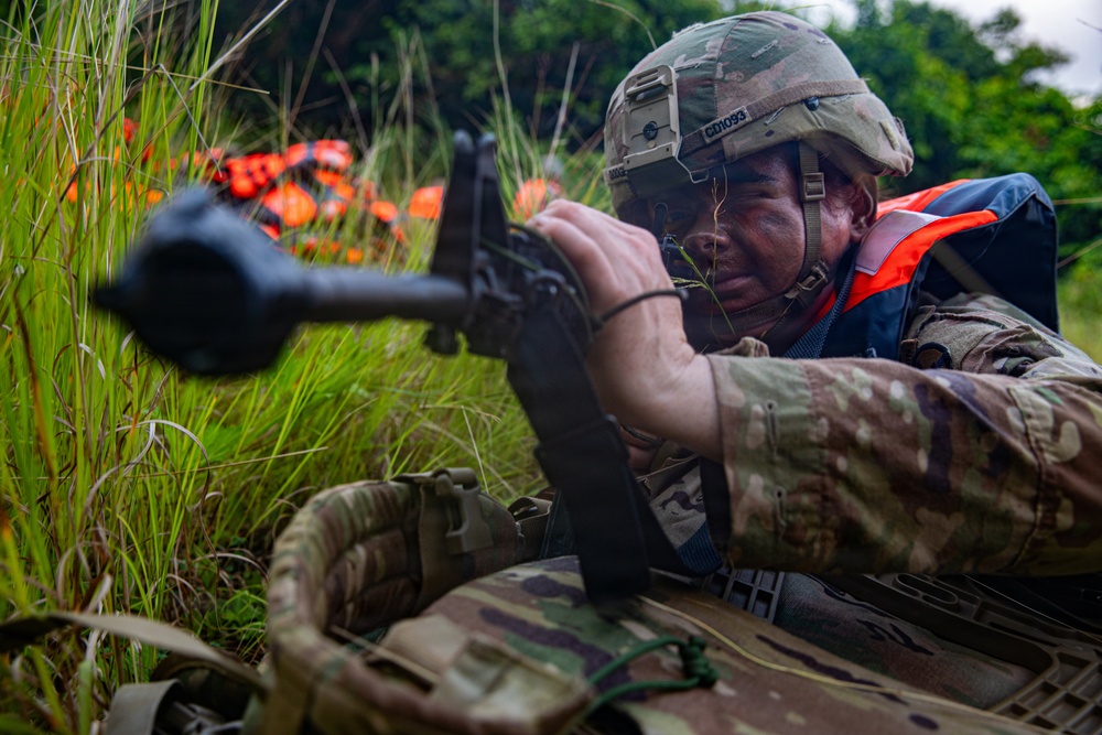 A soldier in full camouflage uniform and tactical helmet is in a prone or low-crouch position aiming a rifle through tall grass. The soldier is wearing an orange life vest over their combat gear, suggesting a water crossing or amphibious training exercise. Additional orange life vests are visible in the background among the vegetation.