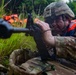 A soldier in full camouflage uniform and tactical helmet is in a prone or low-crouch position aiming a rifle through tall grass. The soldier is wearing an orange life vest over their combat gear, suggesting a water crossing or amphibious training exercise. Additional orange life vests are visible in the background among the vegetation.