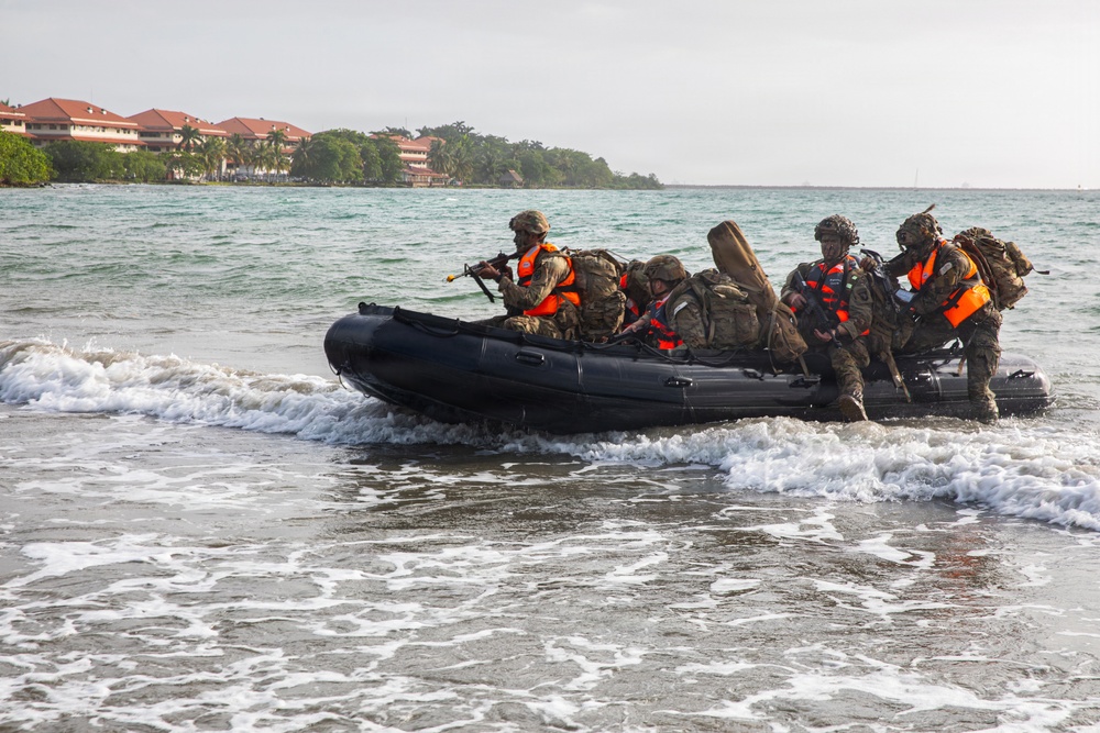 A group of military personnel in camouflage uniforms and orange life vests are arriving at a beach shore aboard a black inflatable rubber raiding craft (CRRC). The soldiers are heavily equipped with tactical backpacks and at least one visible rifle. In the background, a coastal area with palm trees and multi-story buildings with red roofs is visible.