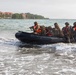 A group of military personnel in camouflage uniforms and orange life vests are arriving at a beach shore aboard a black inflatable rubber raiding craft (CRRC). The soldiers are heavily equipped with tactical backpacks and at least one visible rifle. In the background, a coastal area with palm trees and multi-story buildings with red roofs is visible.