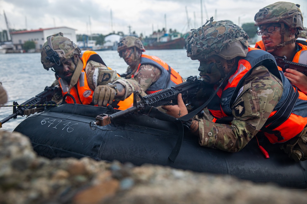 Military personnel in combat gear are conducting an amphibious training exercise from a black inflatable rubber assault craft (CRRC) marked '64146'. The soldiers are wearing multicam uniforms, helmets with camouflage netting, and bright orange life vests, and are armed with M4-style rifles. The background shows a harbor or port environment with vessels and tropical vegetation visible.