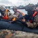 Military personnel in combat gear are conducting an amphibious training exercise from a black inflatable rubber assault craft (CRRC) marked '64146'. The soldiers are wearing multicam uniforms, helmets with camouflage netting, and bright orange life vests, and are armed with M4-style rifles. The background shows a harbor or port environment with vessels and tropical vegetation visible.