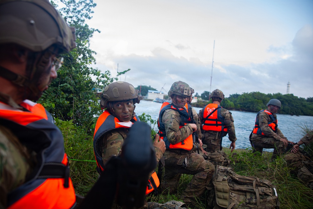 A group of military personnel in camouflage uniforms and tactical helmets are positioned along a vegetated shoreline wearing bright orange life vests. They appear to be conducting a waterborne training exercise or amphibious operation near a body of water with port infrastructure visible in the background. The soldiers are in a crouched, alert posture suggesting a tactical insertion or reconnaissance scenario.