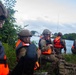 A group of military personnel in camouflage uniforms and tactical helmets are positioned along a vegetated shoreline wearing bright orange life vests. They appear to be conducting a waterborne training exercise or amphibious operation near a body of water with port infrastructure visible in the background. The soldiers are in a crouched, alert posture suggesting a tactical insertion or reconnaissance scenario.