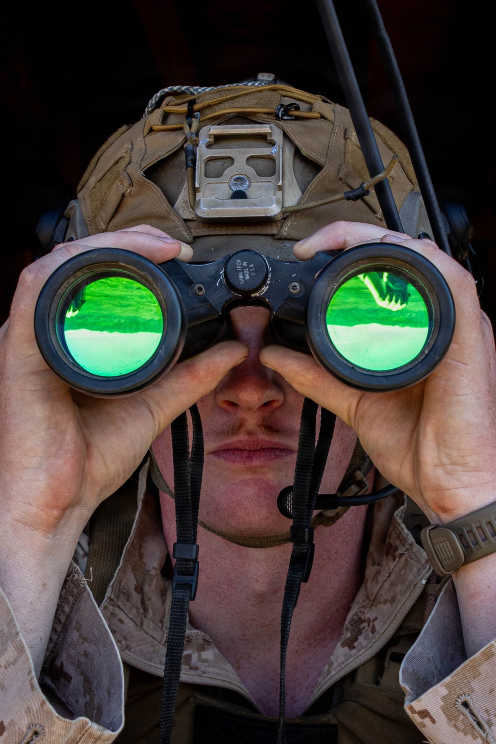 A military service member is looking through binoculars in a close-up frontal photograph. The individual is wearing desert camouflage (MARPAT pattern) uniform and a tan tactical helmet with a night-vision device mount. The binoculars display green-tinted anti-reflective coated lenses and are marked 'KAMA TECH MADE IN USA'.