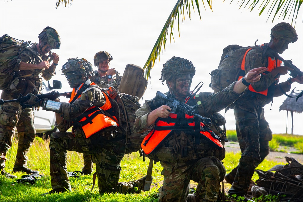 A group of military personnel in camouflage uniforms and tactical gear are conducting what appears to be an amphibious training exercise near a coastal area. Several soldiers are wearing bright orange life vests over their combat equipment, indicating a water-based operation. The soldiers are armed with assault rifles and carrying heavy rucksacks, with one soldier appearing to give hand signals.