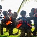 A group of military personnel in camouflage uniforms and tactical gear are conducting what appears to be an amphibious training exercise near a coastal area. Several soldiers are wearing bright orange life vests over their combat equipment, indicating a water-based operation. The soldiers are armed with assault rifles and carrying heavy rucksacks, with one soldier appearing to give hand signals.