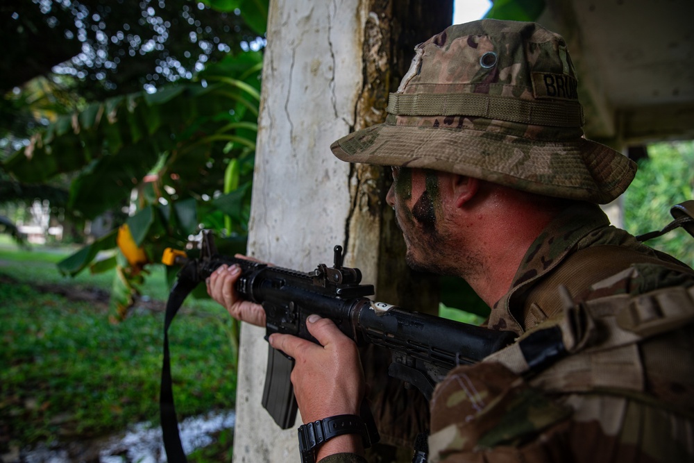 A soldier in MultiCam/OCP camouflage uniform is taking a tactical position behind a concrete pillar or wall, aiming a rifle in a lush, tropical or subtropical environment. The soldier is wearing a boonie hat and has face paint applied, suggesting a field training exercise or operational deployment. A partial name tape reading 'BRO...' is visible on the uniform.