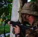 A soldier in MultiCam/OCP camouflage uniform is taking a tactical position behind a concrete pillar or wall, aiming a rifle in a lush, tropical or subtropical environment. The soldier is wearing a boonie hat and has face paint applied, suggesting a field training exercise or operational deployment. A partial name tape reading 'BRO...' is visible on the uniform.