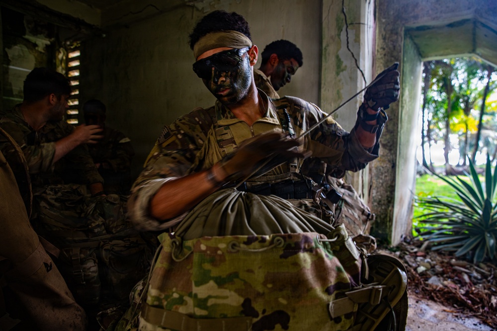 Military personnel in camouflage uniforms with face paint are inside a deteriorating concrete structure, appearing to prepare or stage equipment. The central figure is applying or adjusting face camouflage paint while handling gear, with a large pack visible in the foreground. The setting appears to be a training exercise in a tropical environment based on the vegetation visible through the opening.