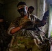 Military personnel in camouflage uniforms with face paint are inside a deteriorating concrete structure, appearing to prepare or stage equipment. The central figure is applying or adjusting face camouflage paint while handling gear, with a large pack visible in the foreground. The setting appears to be a training exercise in a tropical environment based on the vegetation visible through the opening.