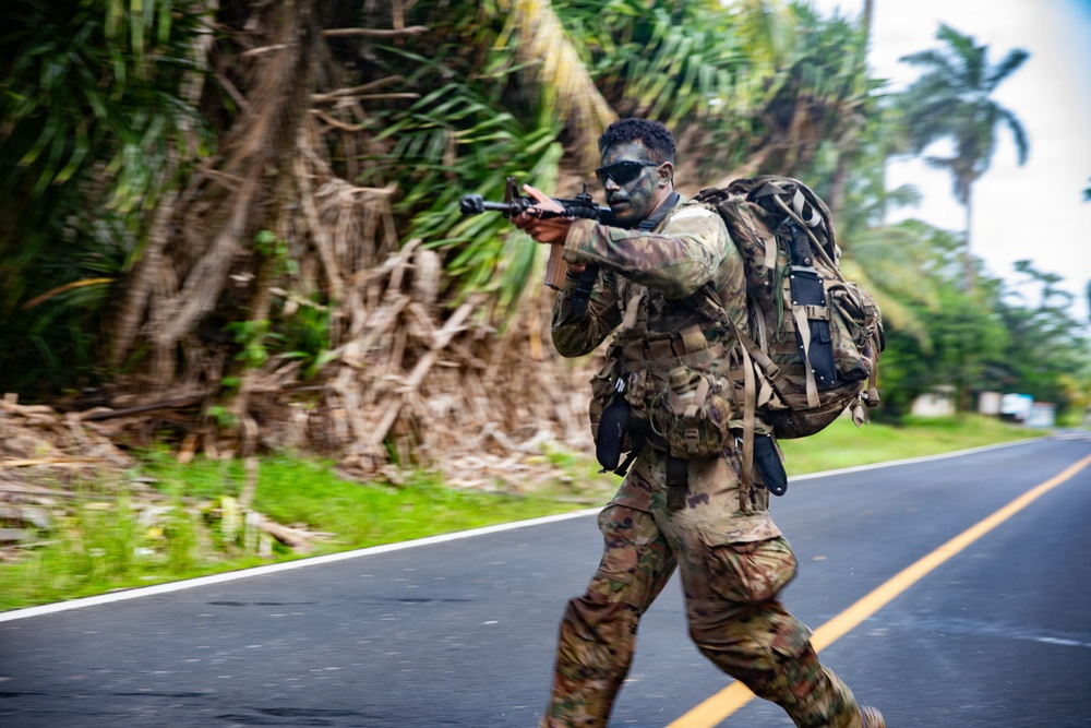 A soldier in full combat gear is moving tactically across a paved road in what appears to be a tropical environment. The individual is wearing camouflage uniform with face paint, carrying a large rucksack, and aiming a rifle while in motion. The background features dense tropical vegetation including palm trees, suggesting a training exercise or operational movement in a Pacific or Caribbean-type region.