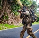 A soldier in full combat gear is moving tactically across a paved road in what appears to be a tropical environment. The individual is wearing camouflage uniform with face paint, carrying a large rucksack, and aiming a rifle while in motion. The background features dense tropical vegetation including palm trees, suggesting a training exercise or operational movement in a Pacific or Caribbean-type region.