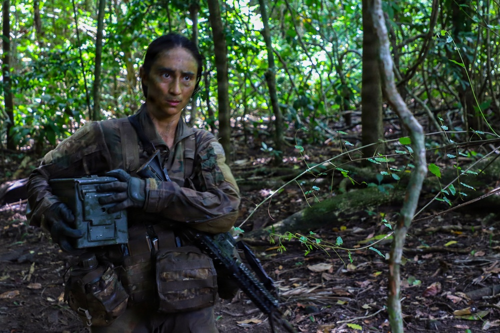 A female soldier in camouflage uniform is positioned in a dense tropical forest, carrying military equipment including a rifle and what appears to be an ammunition box or equipment case. Her face has tactical camouflage paint applied, and she is wearing black tactical gloves. The setting appears to be a jungle training exercise or operational patrol.