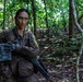 A female soldier in camouflage uniform is positioned in a dense tropical forest, carrying military equipment including a rifle and what appears to be an ammunition box or equipment case. Her face has tactical camouflage paint applied, and she is wearing black tactical gloves. The setting appears to be a jungle training exercise or operational patrol.