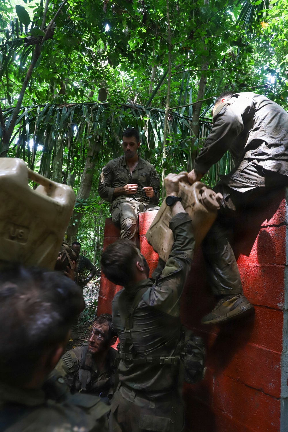 A group of military personnel in olive/camouflage uniforms are conducting what appears to be a training exercise in a jungle or tropical forest environment. They are navigating or scaling a red-painted concrete block wall, with some soldiers boosting others over the obstacle. The soldiers appear muddy and fatigued, consistent with an endurance or obstacle course training scenario.