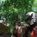 A group of military personnel in olive/camouflage uniforms are conducting what appears to be a training exercise in a jungle or tropical forest environment. They are navigating or scaling a red-painted concrete block wall, with some soldiers boosting others over the obstacle. The soldiers appear muddy and fatigued, consistent with an endurance or obstacle course training scenario.