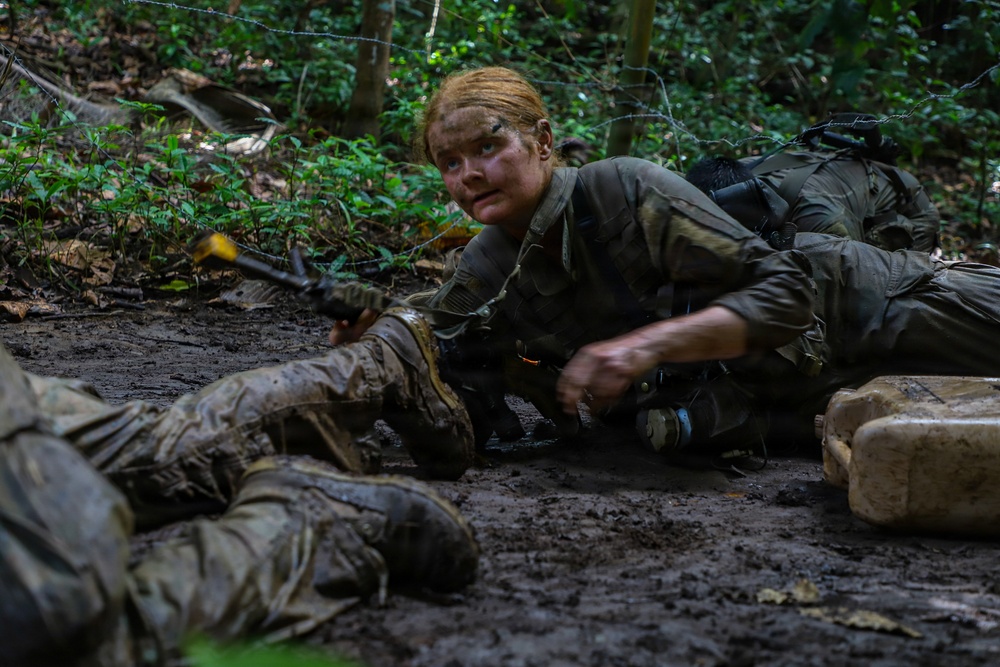 A female soldier in olive drab military uniform is crawling through muddy terrain in a wooded area, appearing to be engaged in a military training exercise. Her face is covered in camouflage paint and mud, and she is holding what appears to be a training weapon with a yellow safety marker. At least one other soldier is visible crawling nearby, and barbed wire can be seen strung above them.