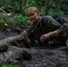 A female soldier in olive drab military uniform is crawling through muddy terrain in a wooded area, appearing to be engaged in a military training exercise. Her face is covered in camouflage paint and mud, and she is holding what appears to be a training weapon with a yellow safety marker. At least one other soldier is visible crawling nearby, and barbed wire can be seen strung above them.
