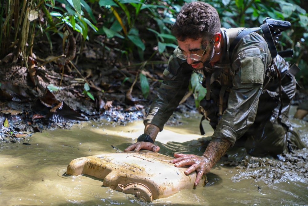 A soldier or military personnel in camouflage uniform, with face paint and glasses, is kneeling in a muddy jungle stream or shallow water. He is handling a dirty, mud-covered plastic container or canister that is partially submerged. The setting appears to be a dense tropical jungle environment during what looks like a training exercise or field operation.