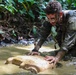 A soldier or military personnel in camouflage uniform, with face paint and glasses, is kneeling in a muddy jungle stream or shallow water. He is handling a dirty, mud-covered plastic container or canister that is partially submerged. The setting appears to be a dense tropical jungle environment during what looks like a training exercise or field operation.