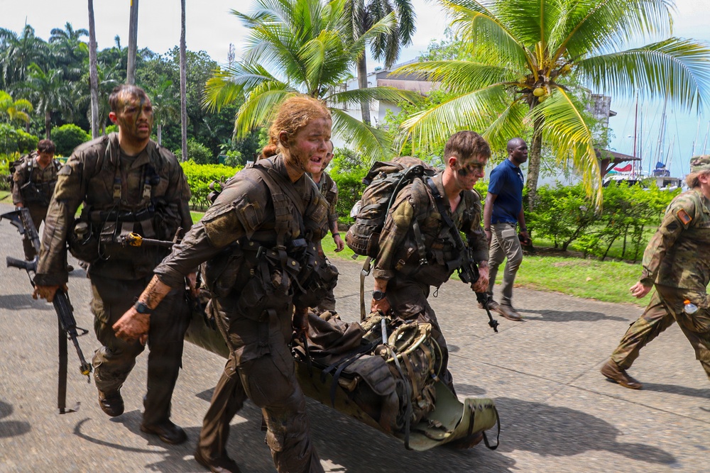 A group of military personnel in full combat gear, with faces painted in camouflage and uniforms covered in mud, are moving urgently along a paved path in a tropical environment. Two soldiers in the foreground are carrying what appears to be a field litter or stretcher, suggesting a casualty evacuation drill or training exercise. The setting features palm trees and tropical vegetation, consistent with a warm-climate training location.