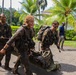 A group of military personnel in full combat gear, with faces painted in camouflage and uniforms covered in mud, are moving urgently along a paved path in a tropical environment. Two soldiers in the foreground are carrying what appears to be a field litter or stretcher, suggesting a casualty evacuation drill or training exercise. The setting features palm trees and tropical vegetation, consistent with a warm-climate training location.