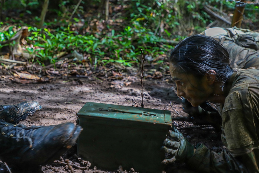 A person in camouflage/military-style clothing is lying prone in muddy terrain within a forested environment. They are closely examining or interacting with a green metal ammunition box or military storage container. The individual appears heavily soiled with mud, suggesting prolonged activity in wet, jungle-like conditions.