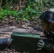 A person in camouflage/military-style clothing is lying prone in muddy terrain within a forested environment. They are closely examining or interacting with a green metal ammunition box or military storage container. The individual appears heavily soiled with mud, suggesting prolonged activity in wet, jungle-like conditions.