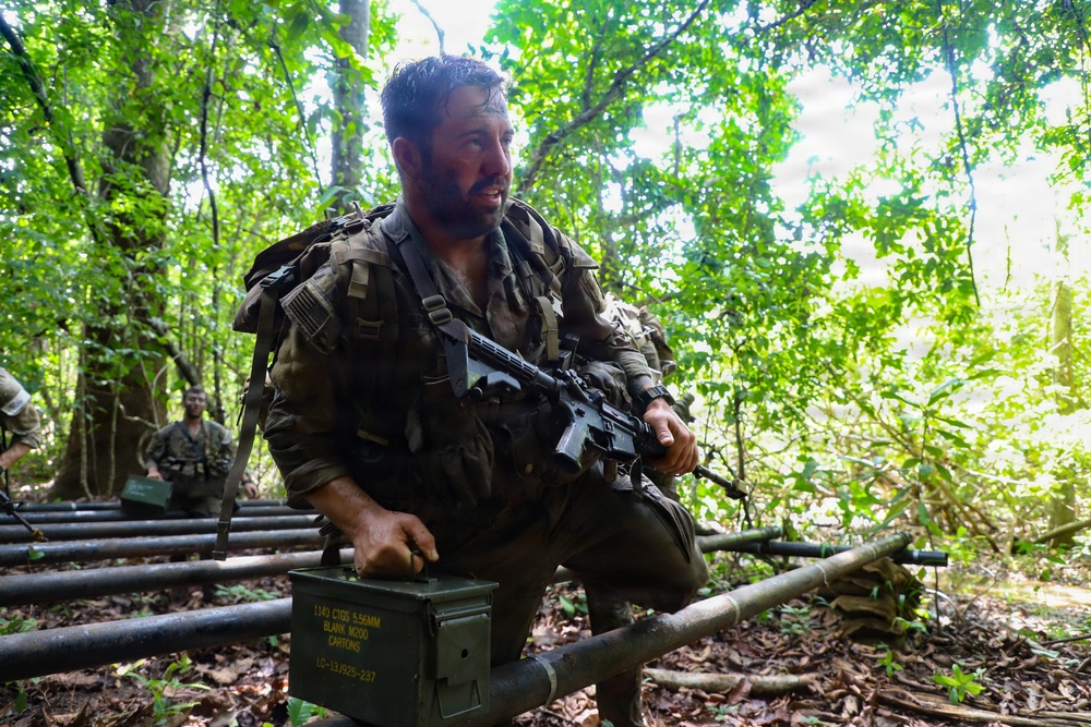 A heavily equipped military personnel is crouching in a jungle/forest environment, holding an assault rifle and resting a hand on a green military ammunition box. The soldier appears wet and fatigued, wearing full tactical gear including a backpack and combat uniform. Additional personnel are visible in the background, suggesting a group training exercise or field operation.