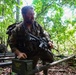 A heavily equipped military personnel is crouching in a jungle/forest environment, holding an assault rifle and resting a hand on a green military ammunition box. The soldier appears wet and fatigued, wearing full tactical gear including a backpack and combat uniform. Additional personnel are visible in the background, suggesting a group training exercise or field operation.
