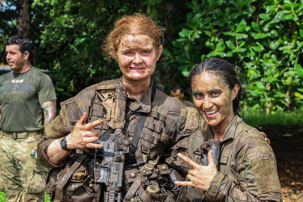 Two female soldiers in full tactical gear and camouflage uniforms are posing together, both heavily covered in mud, suggesting completion of a demanding field training exercise. They are smiling and making hand gestures, indicating a celebratory or triumphant mood. A third individual in a green 'JUNGLE' t-shirt and camouflage pants is visible in the background.
