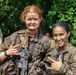 Two female soldiers in full tactical gear and camouflage uniforms are posing together, both heavily covered in mud, suggesting completion of a demanding field training exercise. They are smiling and making hand gestures, indicating a celebratory or triumphant mood. A third individual in a green 'JUNGLE' t-shirt and camouflage pants is visible in the background.