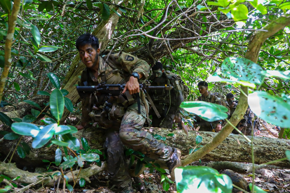 Military personnel in full combat gear are navigating through dense tropical jungle terrain, climbing over fallen logs and moving through thick vegetation. The lead soldier is prominently visible carrying an assault rifle while wearing multicam camouflage uniform with tactical vest and backpack. At least two additional soldiers are visible in the background following the same route through the forest.