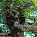 Military personnel in full combat gear are navigating through dense tropical jungle terrain, climbing over fallen logs and moving through thick vegetation. The lead soldier is prominently visible carrying an assault rifle while wearing multicam camouflage uniform with tactical vest and backpack. At least two additional soldiers are visible in the background following the same route through the forest.