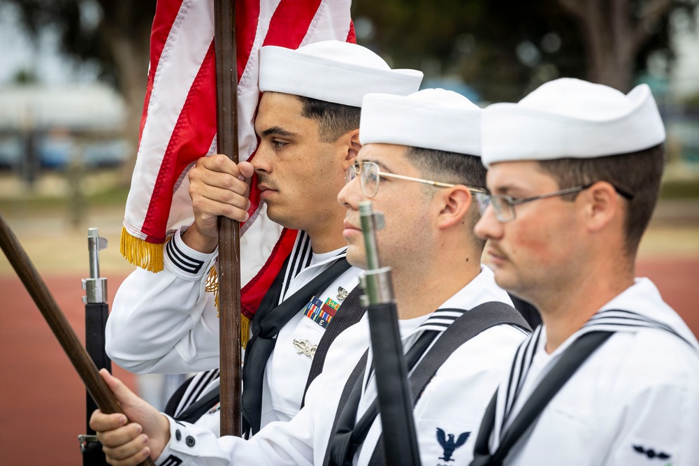 Three U.S. Navy sailors in dress white uniforms stand in formation as part of a color guard ceremony. The lead sailor is gripping the staff of an American flag with gold fringe, while the others carry rifles at their sides. The scene appears to be an outdoor military ceremonial event.