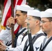 Three U.S. Navy sailors in dress white uniforms stand in formation as part of a color guard ceremony. The lead sailor is gripping the staff of an American flag with gold fringe, while the others carry rifles at their sides. The scene appears to be an outdoor military ceremonial event.