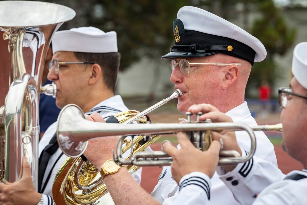 A U.S. Navy military band is performing outdoors, with multiple musicians in dress white uniforms playing brass instruments. The central figure, wearing a senior enlisted officer's combination cover with gold insignia, is actively playing a silver trumpet or flugelhorn. The setting appears to be a parade ground or athletic field based on the visible track surface in the background.