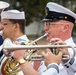 A U.S. Navy military band is performing outdoors, with multiple musicians in dress white uniforms playing brass instruments. The central figure, wearing a senior enlisted officer's combination cover with gold insignia, is actively playing a silver trumpet or flugelhorn. The setting appears to be a parade ground or athletic field based on the visible track surface in the background.