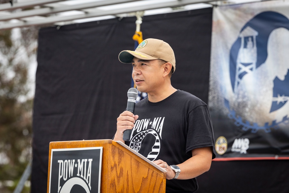 A man is speaking at a podium during a POW-MIA commemorative event. He is wearing a black POW-MIA t-shirt and a tan baseball cap while holding a microphone. The backdrop features the iconic POW-MIA flag imagery on a black banner.