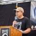 A man is speaking at a podium during a POW-MIA commemorative event. He is wearing a black POW-MIA t-shirt and a tan baseball cap while holding a microphone. The backdrop features the iconic POW-MIA flag imagery on a black banner.