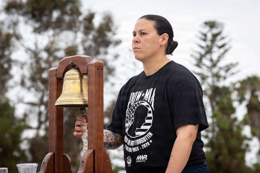 A person wearing a black POW/MIA commemorative t-shirt stands solemnly beside a brass bell mounted on a wooden frame. The individual holds the bell's rope clapper, suggesting participation in a memorial or remembrance ceremony. The setting appears to be an outdoor event with trees visible in the background.