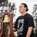 A person wearing a black POW/MIA commemorative t-shirt stands solemnly beside a brass bell mounted on a wooden frame. The individual holds the bell's rope clapper, suggesting participation in a memorial or remembrance ceremony. The setting appears to be an outdoor event with trees visible in the background.