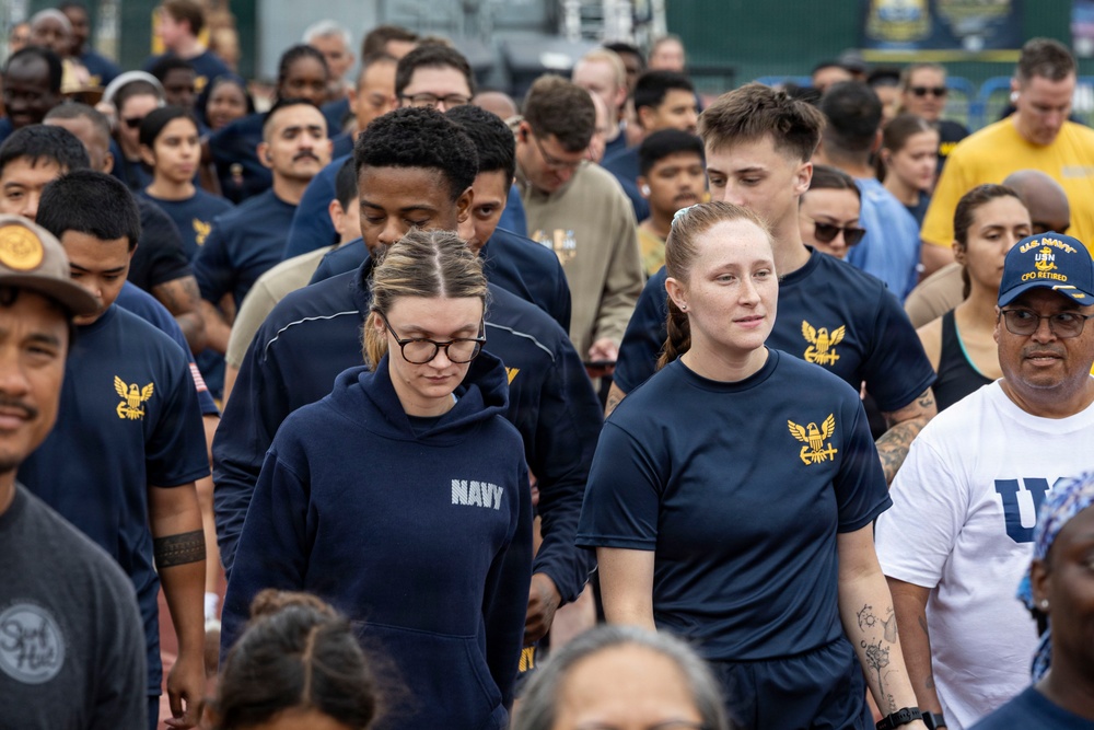 A large group of U.S. Navy personnel are participating in what appears to be a group walk or physical fitness event. The majority are wearing standard Navy physical training (PT) gear including navy blue t-shirts and hoodies with gold eagle insignia. A mix of active duty members and at least one retired CPO (Chief Petty Officer) are visible in the crowd.